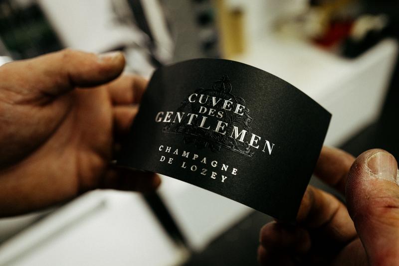 Two hands holding a Champagne De Lozey Cuvée des Gentlemen label at print shop