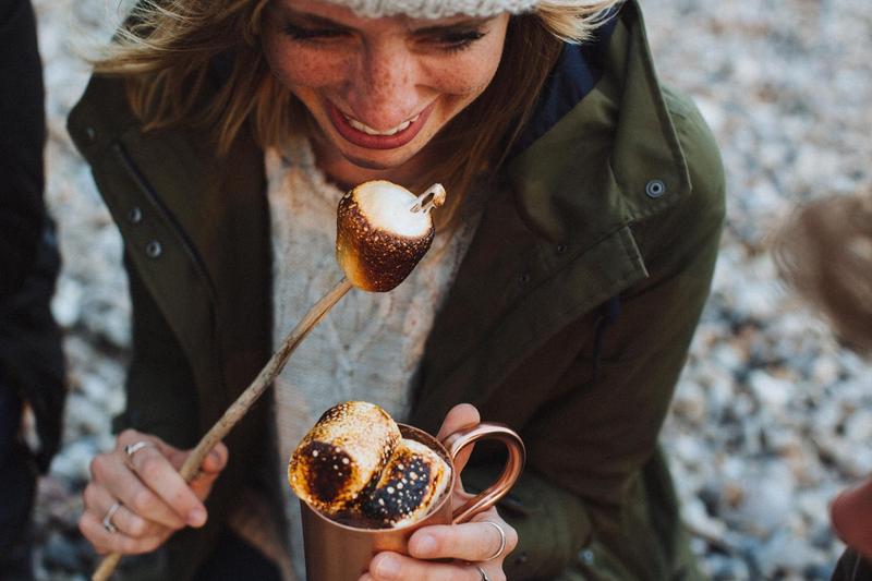 Close up of a girl in winter clothes eating a campfire marshmallow on a stick and holding a mug of hot chocolate