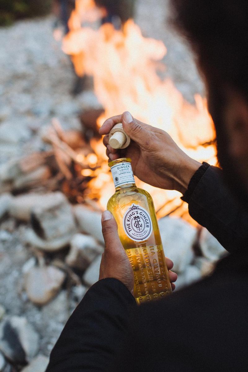 Close up of man opening a bottle of The King's Ginger with a campfire in the background