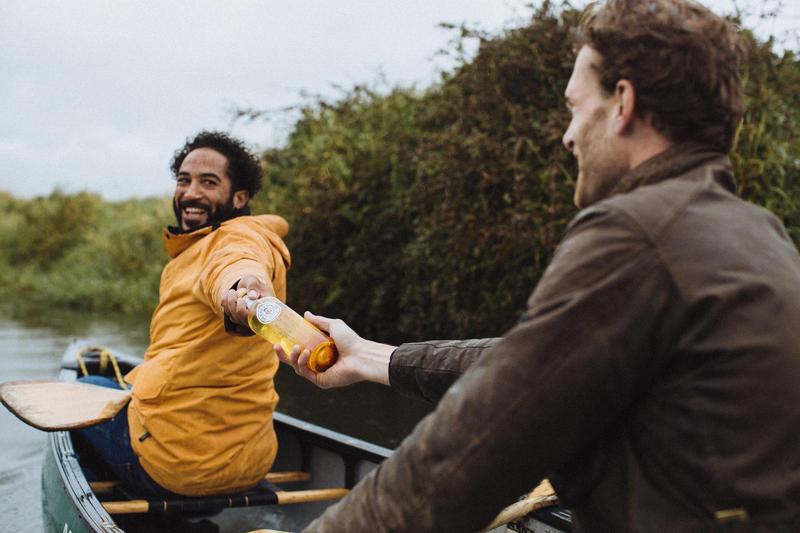 One man hands a bottle of The King's Ginger to another sitting in a canoe