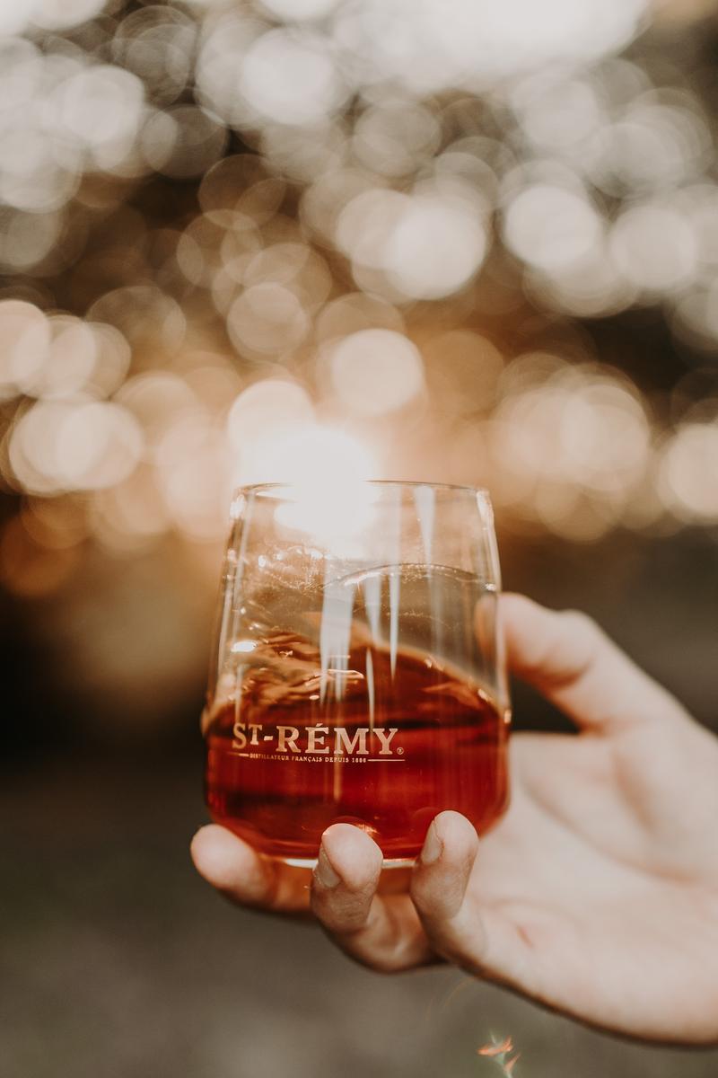 Close up of hand holding glass of St-Rémy brandy in front of sunset through trees